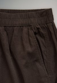 Gros plan d'un pantalon en tissu marron foncé avec taille élastique et détail de poche latérale.