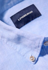 Ljusblå linneskjorta med krage, en enda knappstängning och en marinblå etikett som visar "LINDBERGH" i guldtext.