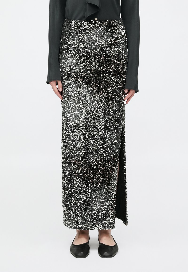 Gonna maxi nera in paillettes con uno spacco laterale, caratterizzata da una texture lucida e una silhouette aderente. Abbinata a ballerine nere.