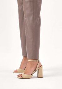 Beżowe sandały na obcasie z odkrytymi palcami, z szerokim obcasem blokowym i paskiem wokół kostki, zestawione z dopasowanymi, jasnobrązowymi skórzanymi spodniami. Gładka faktura i minimalistyczny design.