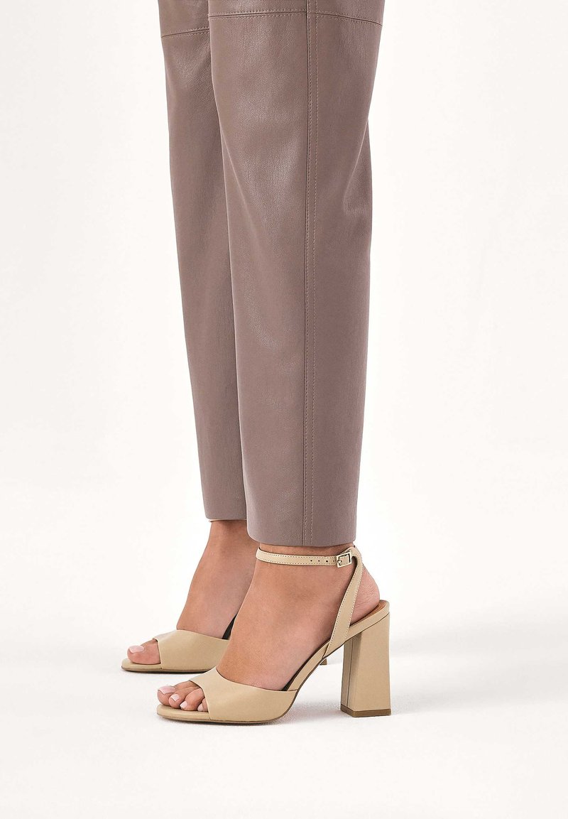 Beżowe sandały na obcasie z odkrytymi palcami, z szerokim obcasem blokowym i paskiem wokół kostki, zestawione z dopasowanymi, jasnobrązowymi skórzanymi spodniami. Gładka faktura i minimalistyczny design.