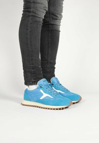 Blue suede sneakers with white accents, round toe, textured panels, and a light rubber sole featuring a serrated edge. Worn with dark jeans.