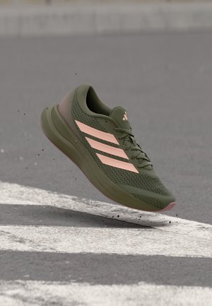 Grüner Adidas Laufschuh mit pinken Streifen und Sohle, der über einer grauen Asphaltstraße mit weißen Markierungen schwebt.