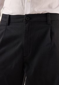 Pantaloni neri realizzati in tessuto intrecciato, dotati di chiusura con bottone e una sola tasca laterale. Texture liscia con vestibilità sartoriale.