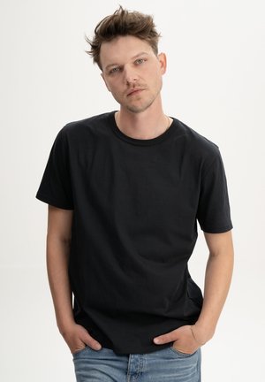 Man met warrig haar, een eenvoudig zwart T-shirt en een blauwe spijkerbroek, staat met zijn handen in zijn zakken tegen een witte achtergrond.