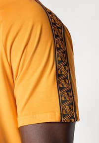 Oranje T-shirt met een zwarte zijstripe met geometrische patronen. Katoenen materiaal, korte mouwen en een ronde hals.