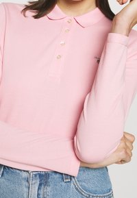 Långärmad rosa polo-tröja med krage och fem knappar, med en texturerad tyg och en liten logotyp på bröstet.