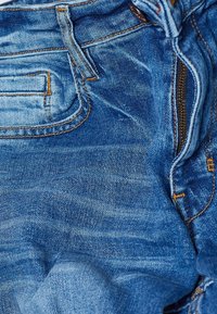 Blå denimjeans med en bleknad, texturerad yta. Har synlig sömnad i orange, en framdragkedja och en bakficka.