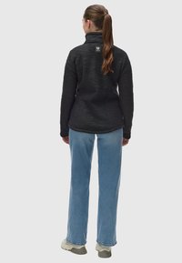 Črna teksturirana jakna s stoječim ovratnikom, ki ima logotip na hrbtni strani, v kombinaciji s svetlo modrimi jeans hlačami in supergami nevtralne barve.