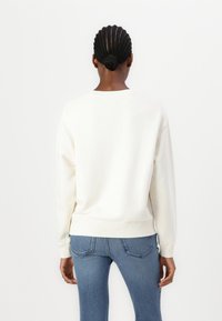 Mikina s okrúhlym výstrihom v off-white farbe s rebrovanými manžetami a lemom. V páre s modrými džínsami, zobrazená zo zadného pohľadu.