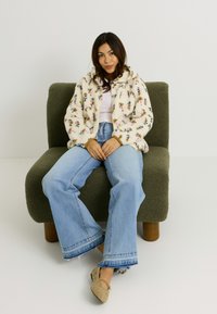Veste en polaire crème à motifs floraux, jean bleu clair décontracté avec ourlet effiloché et chaussures beiges avec embellissements, assis sur une chaise verte texturée.