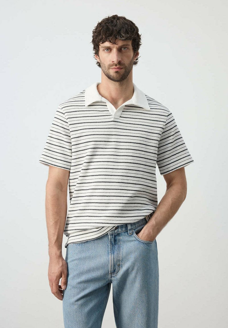 Homme aux cheveux bouclés et foncés, portant un polo blanc à manches courtes avec des rayures noires et un jean bleu clair, debout devant un fond clair uni.