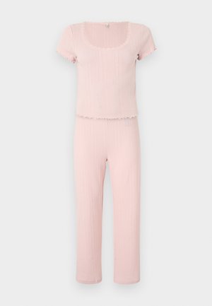 Weiches rosa geripptes Kurzarmoberteil mit gewellten Kanten, kombiniert mit passenden hoch taillierten gerippten Hosen vor schlichtem Hintergrund.