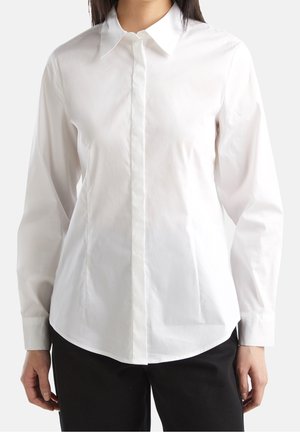 Woman wearing a long-sleeve, buttoned white collared shirt and black pants, shown from shoulders to hips against a light background.