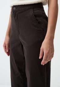 Donkerbruine broek met een gladde textuur, hoge taille, zijzakken en een knoopsluiting. De stof lijkt iets gekreukt.