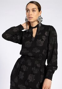 Zwarte jurk met een subtiel bloemenpatroon in metallic draden, met een keyhole-halslijn en een gesmokte taille. Doorzichtige stof en lange mouwen.