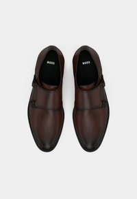 Pāris brūnu ādas monk siksnas kleitu apavu ar dubultiem sprādzēm un melnām zolēm ar uzrakstu "BOSS" uz balta fona.