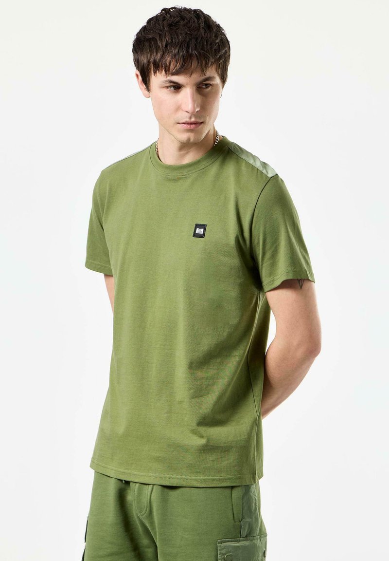Olivenfarvet bomulds T-shirt med korte ærmer, rund halsudskæring og et lille sort logo på brystet. Glat tekstur, let design.