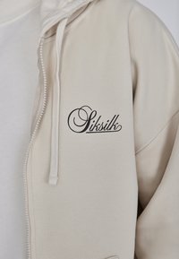 Sudadera beige con cremallera, de textura suave y con cordones, con un distintivo logo negro "Sitsilk" bordado en la zona del pecho.