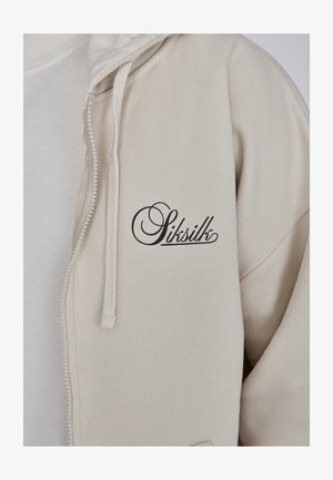 Beige Zip-Hoodie mit weicher Textur und Kordelzügen, mit einem markanten schwarzen Logo "Sitsilk", das auf der Brust gestickt ist.
