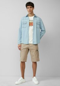 Chemise en denim bleu clair avec poches poitrine, shorts cargo beiges et baskets blanches. Le t-shirt présente une impression graphique d'un paysage.