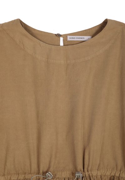 Chemise marron clair avec encolure arrondie et fermeture par bouton au dos, avec un détail élastique froncé près du bord inférieur.