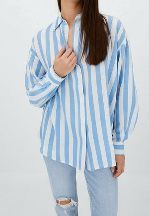 Femme aux longs cheveux bruns, portant une chemise ample bleu et blanc à rayures verticales et un jean déchiré bleu clair, tenant le col de la chemise.
