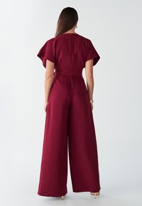 Bordo kombinezon s kratkimi rahlo valovitimi rokavi, pasom, širokimi hlačnicami, gladko teksturo in minimalnimi šivi.