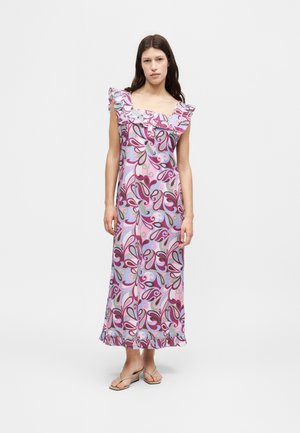 Femme portant une robe sans manches, longue jusqu'aux chevilles, avec des motifs floraux abstraits roses, violets et gris, et un col et un ourlet volantés, debout pieds nus.