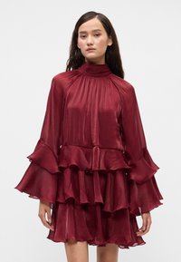 Burgunderfarget, lagdelt kjole med høy hals, lange volangslipper og en utvidet silhuett. Laget av lett, strukturert stoff.