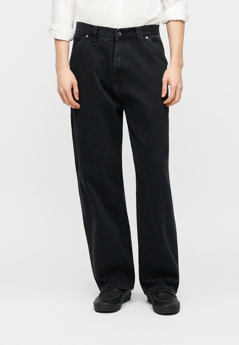 Persona che indossa jeans neri larghi e scarpe nere, con le maniche bianche della camicia arrotolate fino agli avambracci.