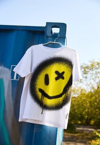 White t-shirt featuring a large yellow smiley face with black spray-painted accents and drips, hanging on a blue container.