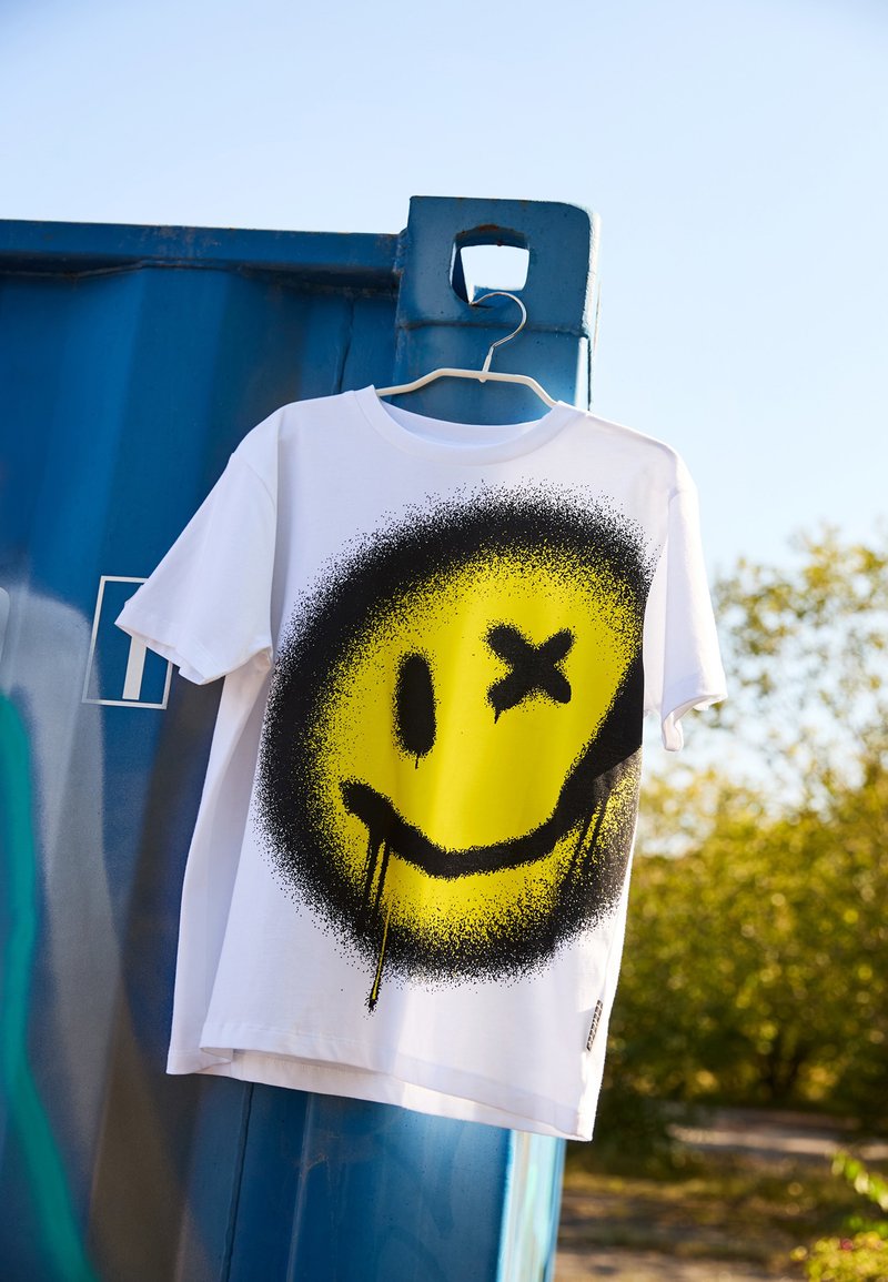 White t-shirt featuring a large yellow smiley face with black spray-painted accents and drips, hanging on a blue container.