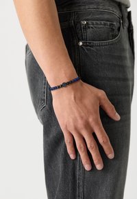 Brățară din mărgele negre care prezintă mărgele rotunde mată de culoare închisă și albastre vibrante, cu un detaliu emblematic central, purtată pe o mână împotriva unor blugi gri.