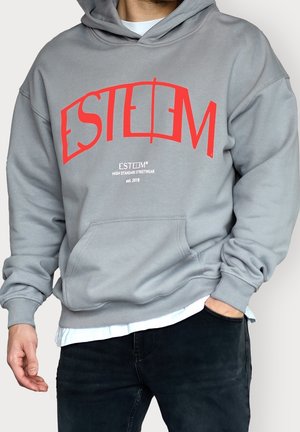 Man met een grijze hoodie met grote rode tekst "ESTEEM" en kleinere witte tekst "High Standard Streetwear est. 2018," gecombineerd met een zwarte broek.