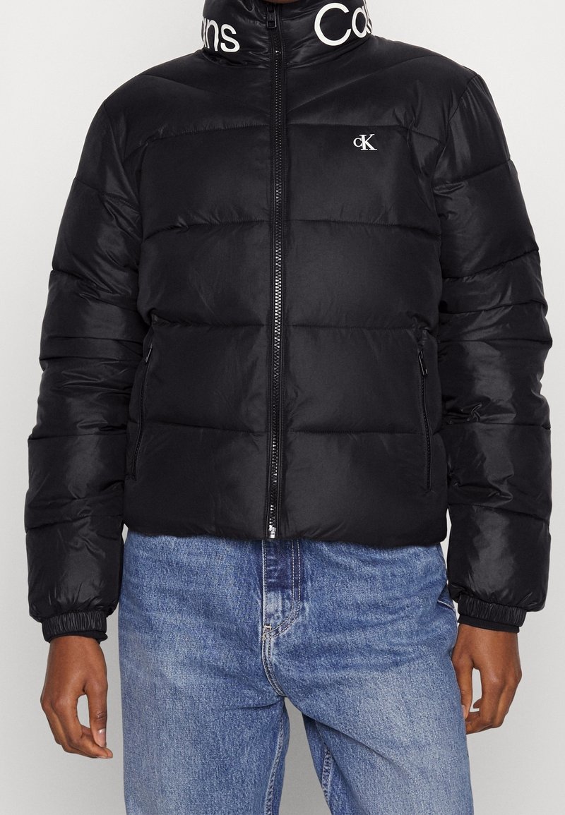 Chaqueta acolchada negra con cierre de cremallera frontal, puños acanalados y cuello alto con logotipo. Presenta un diseño acolchado y una tela elegante y brillante.