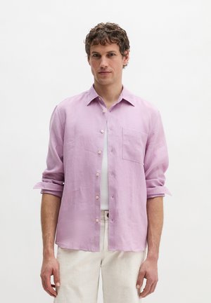 Mand med krøllet hår iført en lys lilla skjorte med knapper over en hvid undertrøje og lyse bukser, stående foran en ensfarvet baggrund.