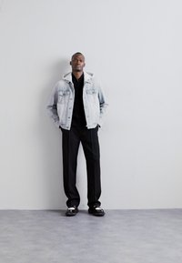 Jeansjack met een grijze vervaging, zwarte trui eronder, slim fit zwarte broeken en zwarte schoenen met witte accenten. Eenvoudige, moderne pasvorm.