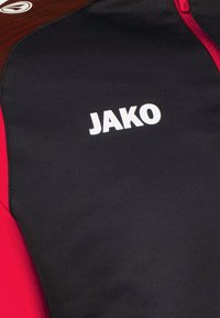 Schwarze und rote Jacke mit einem strukturierten oberen Bereich, auffälligem weißen "JAKO"-Logo und einem durchgehenden Reißverschluss auf der Vorderseite.