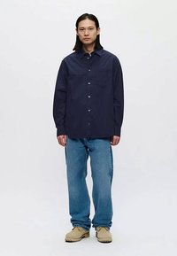 Moški v temno modri srajci z dolgimi rokavi, ohlapnih modrih hlačah in bež čevljih stoji ob preprosti ozadju.