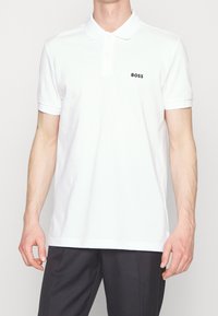 Vit polo med klassisk krage och korta ärmar. Har en liten svart "BOSS"-logotyp på vänster bröst. Tillverkad av mjukt, glatt tyg.