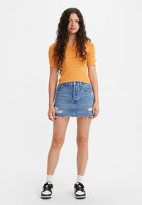 Oranje geribde polo met korte mouwen, gecombineerd met een lichtblauwe denim minirok met een rafelige zoom en versleten accenten. Zwart-witte sneakers.