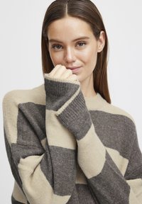 Pull en rayures marron et beige avec une coupe décontractée, texture douce et poignets côtelés. Le design présente de larges rayures horizontales et un col arrondi.