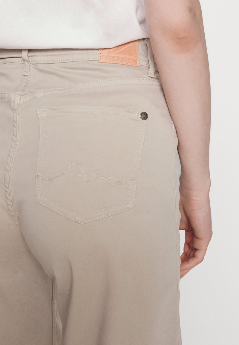 Pantaloni beige dal taglio dritto con una tasca posteriore caratterizzata da cuciture sottili e un bottone metallico. Un'etichetta in pelle arancione è esposta sulla cintura.