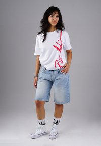 Weißes T-Shirt mit kurzen Ärmeln und rotem diagonalen Text, kombiniert mit hellblauen Jeansshorts. Tragen mit weißen High-Top-Sneakern und gemusterten Socken.
