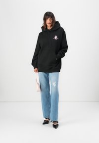 Black hoodie with a front pocket, drawstrings, and colorful embroidered design. Paired with light blue distressed jeans and black pointed shoes.
