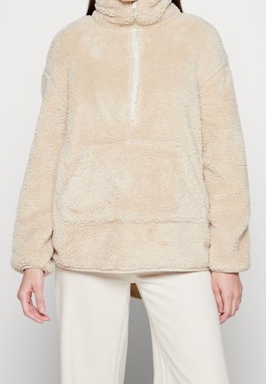 Pull polaire beige duveteux avec demi-fermeture éclair et poche avant, porté avec un pantalon blanc cassé, visible du cou aux hanches.