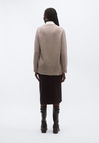 Beež jahe, veidi üle suuruse lõikega, soonikkoes äärega, kantud pruuni põlvikupikkuse seeliku ja põlvikute kohal olevate saapadega, vaadatuna tagant.