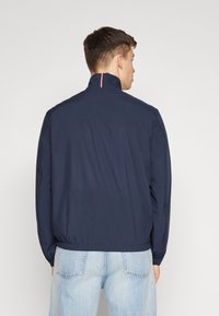 Giacca a vento blu navy con colletto alto, vita elastica e dettagli a strisce rosso e bianco a contrasto sul colletto. Tessuto leggero.