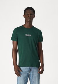 T-shirt verde in cotone con maniche corte, scollatura rotonda e logo bianco "HILFIGER" sul petto. Indossato con jeans azzurri chiari.